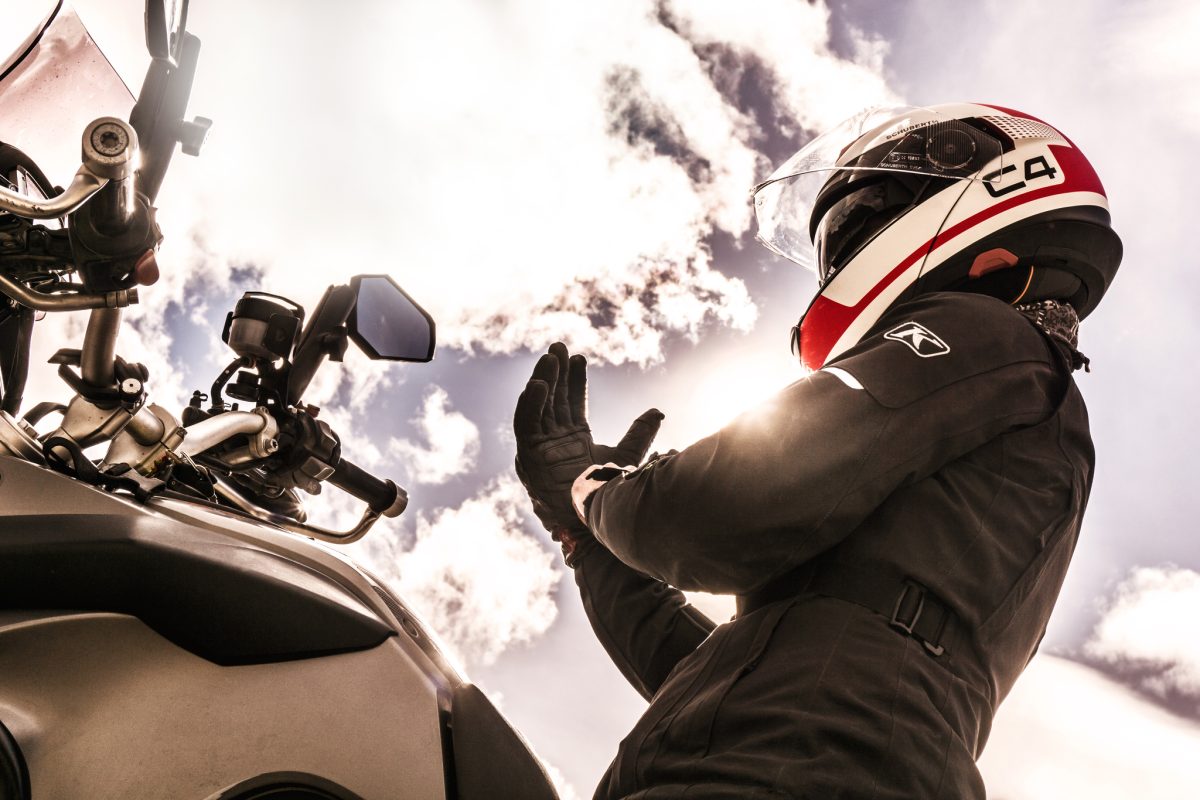 Motard équipé d'un blouson et de gants de protection levé les mains sur sa moto, casque intégral blanc rouge et noir, à contre-jour